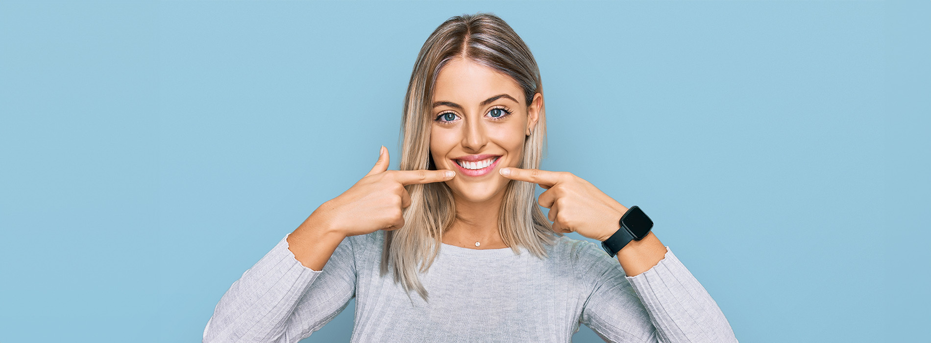 The image is a photograph of a woman with light skin, smiling at the camera. She appears to be in her late twenties or early thirties and has long hair. Her eyes are looking directly at the camera, and she is holding up her index finger near her mouth as if she s making a point or emphasizing something. The background is plain and light-colored, which suggests that this could be a stock photo used for various purposes such as advertising, personal branding, or lifestyle content.