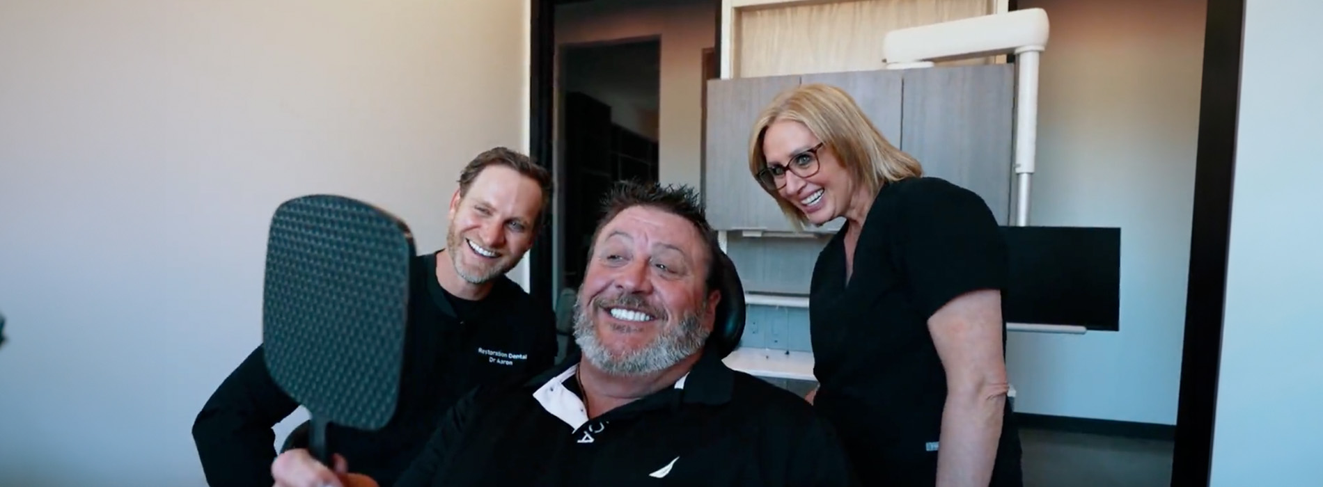 A person is seated in a dental chair, receiving care from a dental professional who stands behind them.